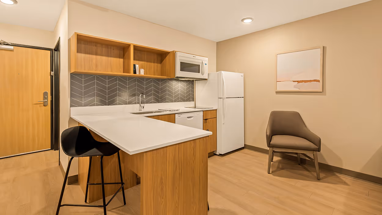 Compact kitchen with wooden cabinets, white countertop, white appliances, a black bar stool, and a gray chair under abstract wall art.