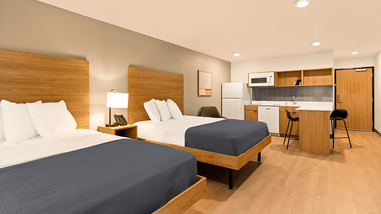 Modern hotel room with two queen beds, wooden furniture, a kitchenette with appliances, and two bar stools.