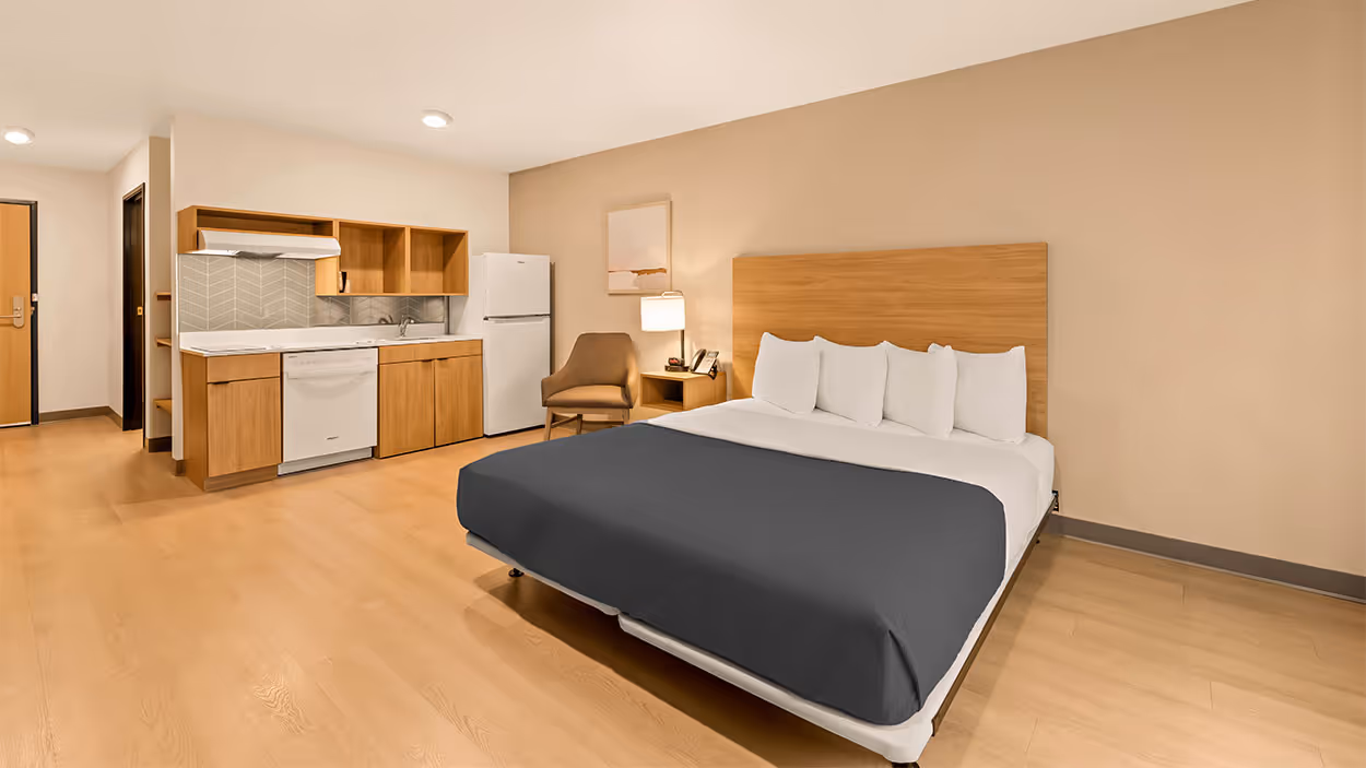 Spacious hotel room with a king bed, wooden headboard, a kitchenette with wooden cabinets, refrigerator, dishwasher, and a brown armchair next to a nightstand with a lamp.