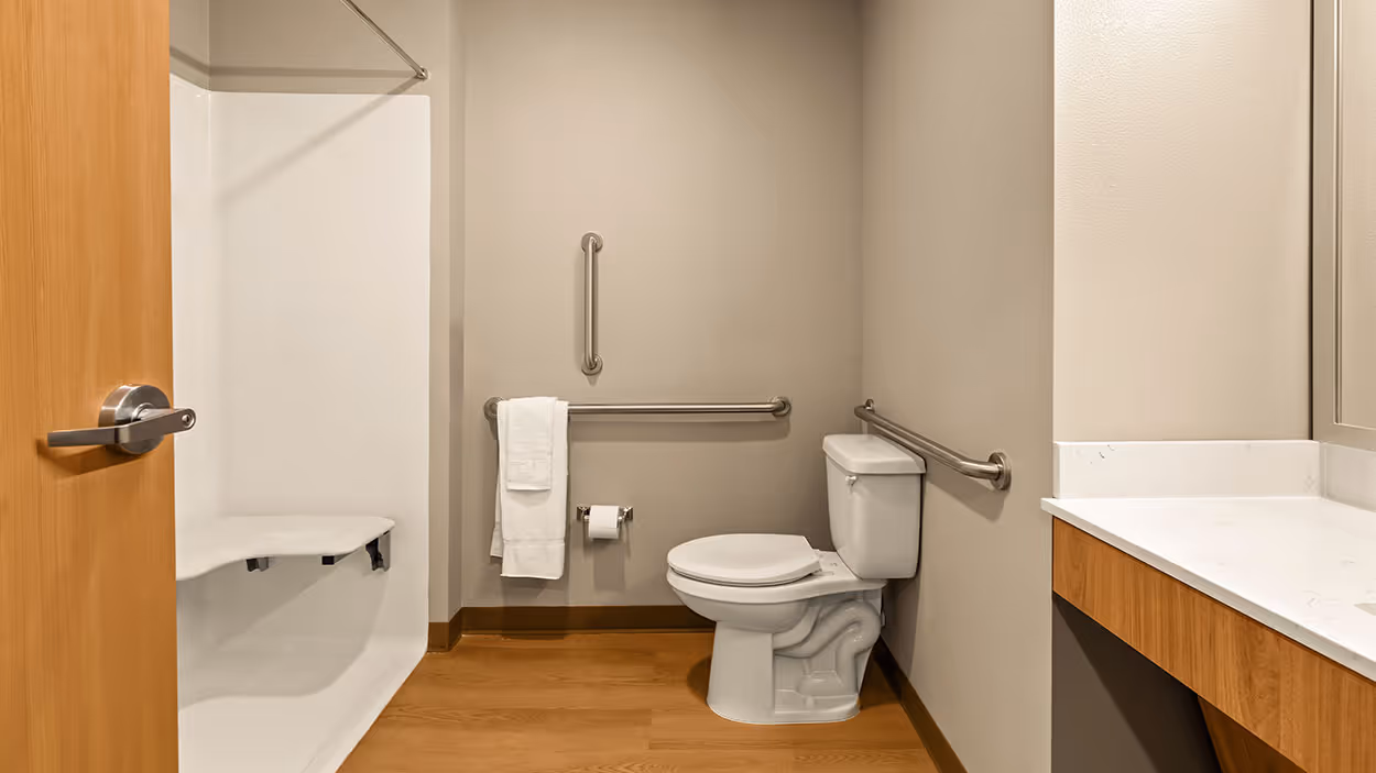 Modern accessible bathroom with white toilet, wall-mounted grab bars, white shower with built-in seat, wooden floor, and wooden vanity with white countertop.