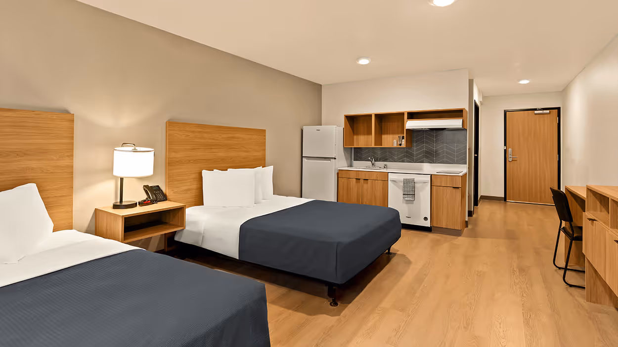 Hotel room with two queen beds with dark gray bedspreads, a nightstand with lamp and phone, a kitchenette with fridge, sink, stove, and dishwasher, and a desk with chair.