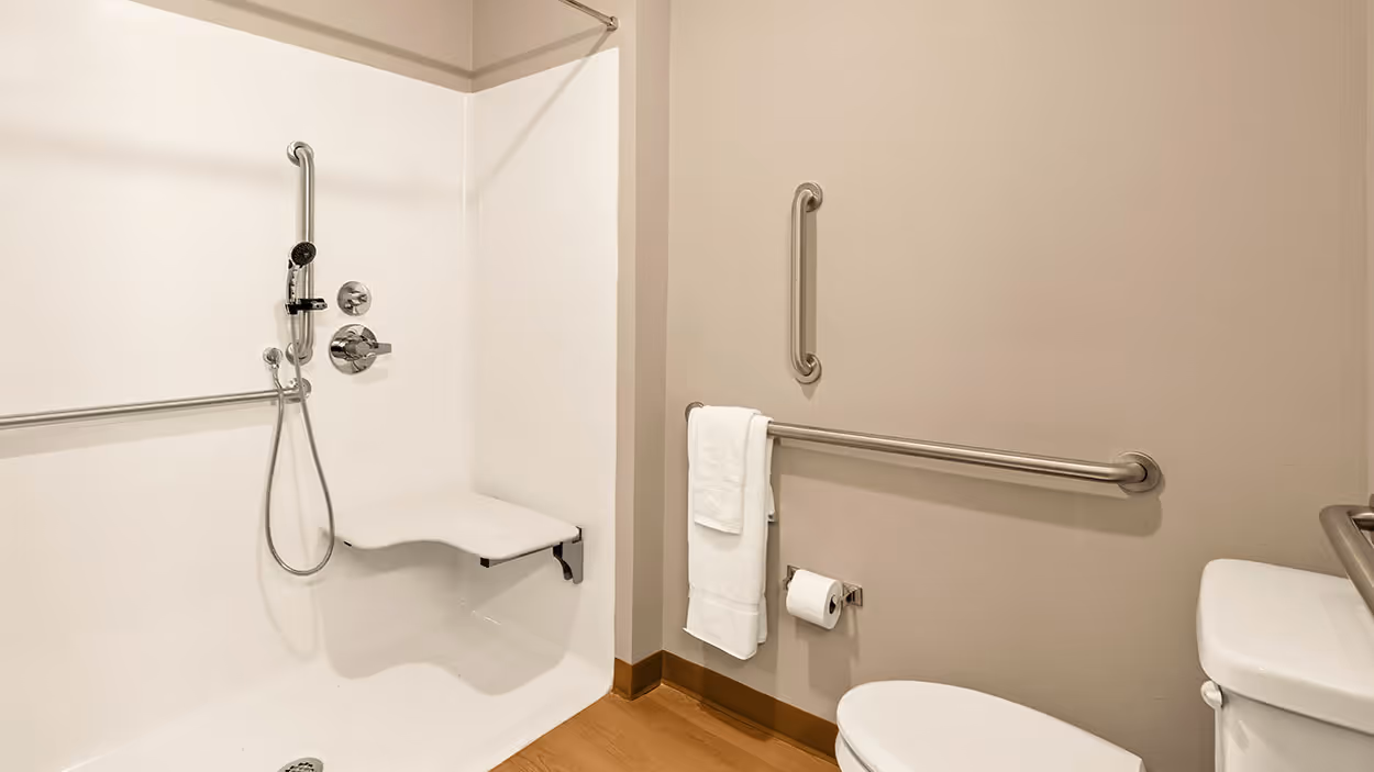 ADA compliant bathroom with white roll-in shower featuring fold-down seat and multiple grab bars, next to toilet with grab bars and towel rack.