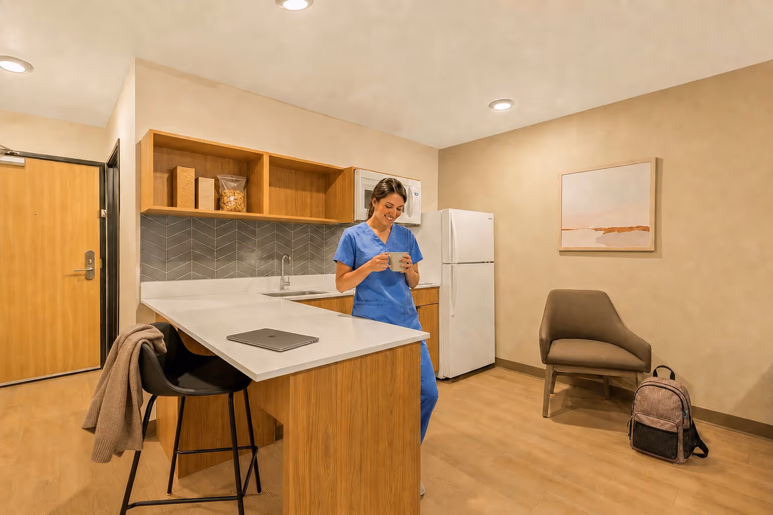 Smiling nurse in blue scrubs standing in a modern kitchen area holding a mug.