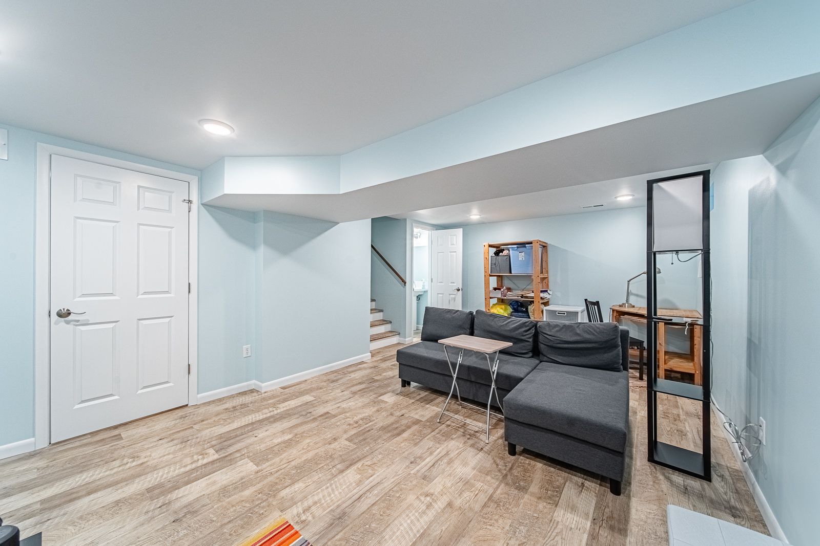 Bright basement media room featuring a gray sectional, desk workspace, and mounted TV across light blue walls.