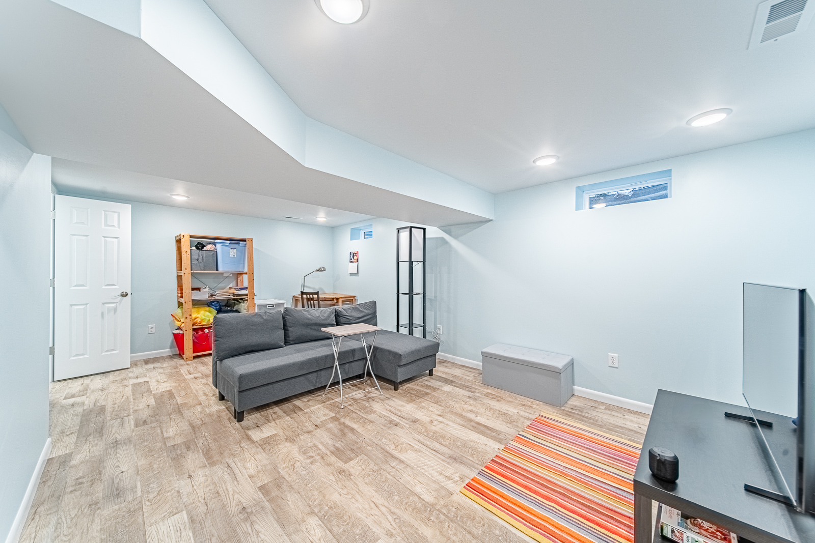 Basement living area with gray sectional, TV, and colorful rug on light wood floors.
