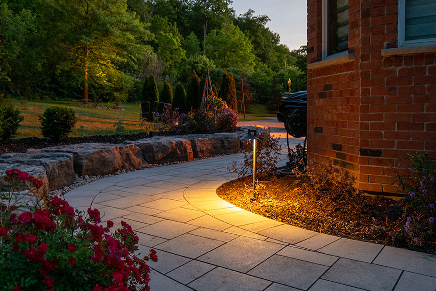Curved stone walkway illuminated by ground lights next to a brick house and garden at dusk.