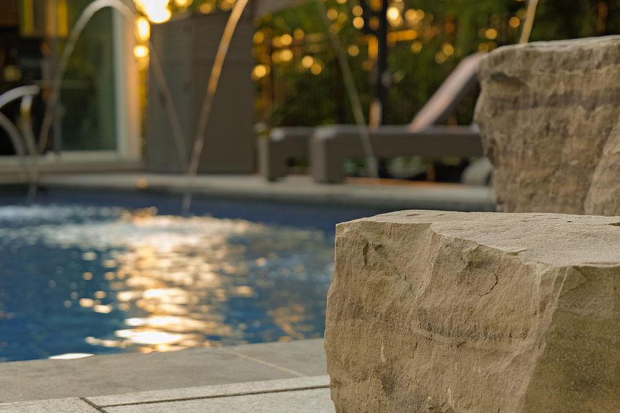 Close-up of textured stone blocks near a pool with water jets and outdoor furniture in the background.