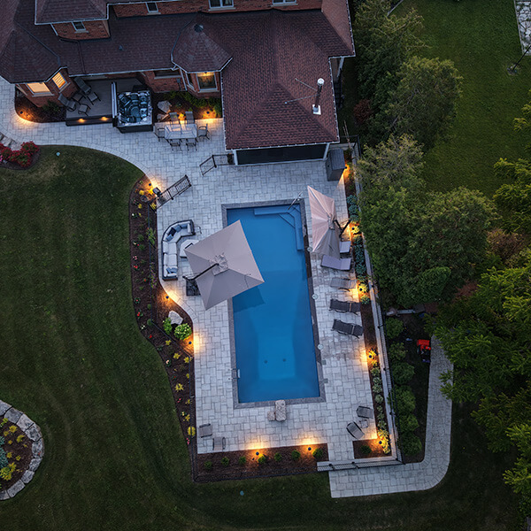 Aerial view of a backyard with a rectangular pool, umbrellas, lounge chairs, and patio lighting at dusk.