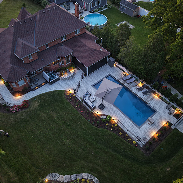Aerial view of a house with a lit patio, rectangular pool with umbrella, surrounded by lawn and trees.