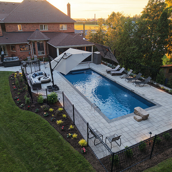 Backyard rectangular swimming pool with lounge chairs, umbrellas, and stone patio near brick house at sunset.