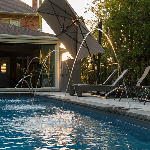 Backyard pool with water jets arching over it and lounge chairs under a large umbrella at sunset.