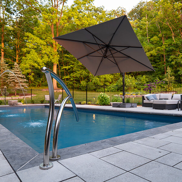 Backyard pool with water jets, large gray umbrella, and outdoor seating area surrounded by green trees.