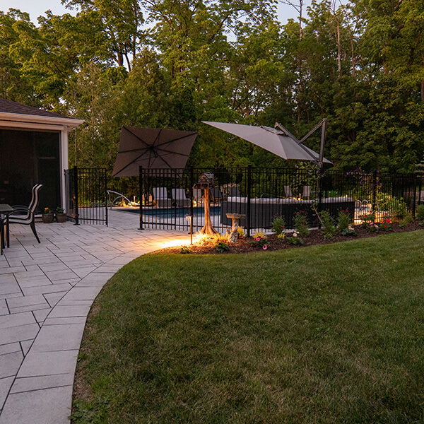 Backyard patio with stone walkway, black fence around pool, umbrellas, and garden lighting at dusk.