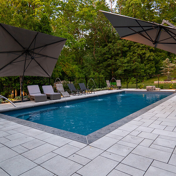 Modern rectangular pool with gray stone deck, lounge chairs, and two large umbrellas in a backyard.