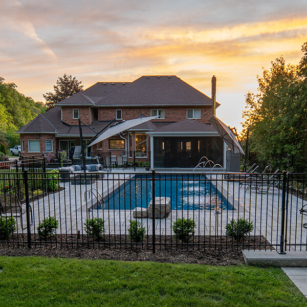 Backyard with a rectangular pool, patio furniture, and a large brick house at sunset.