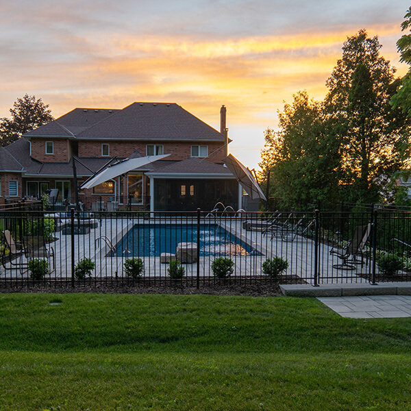 Backyard with a fenced pool, patio seating, and a large brick house during sunset.