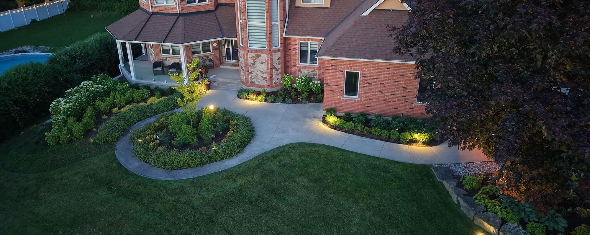 A brick house with landscaped garden beds, curved walkway, green lawn, and outdoor seating on porch at dusk.