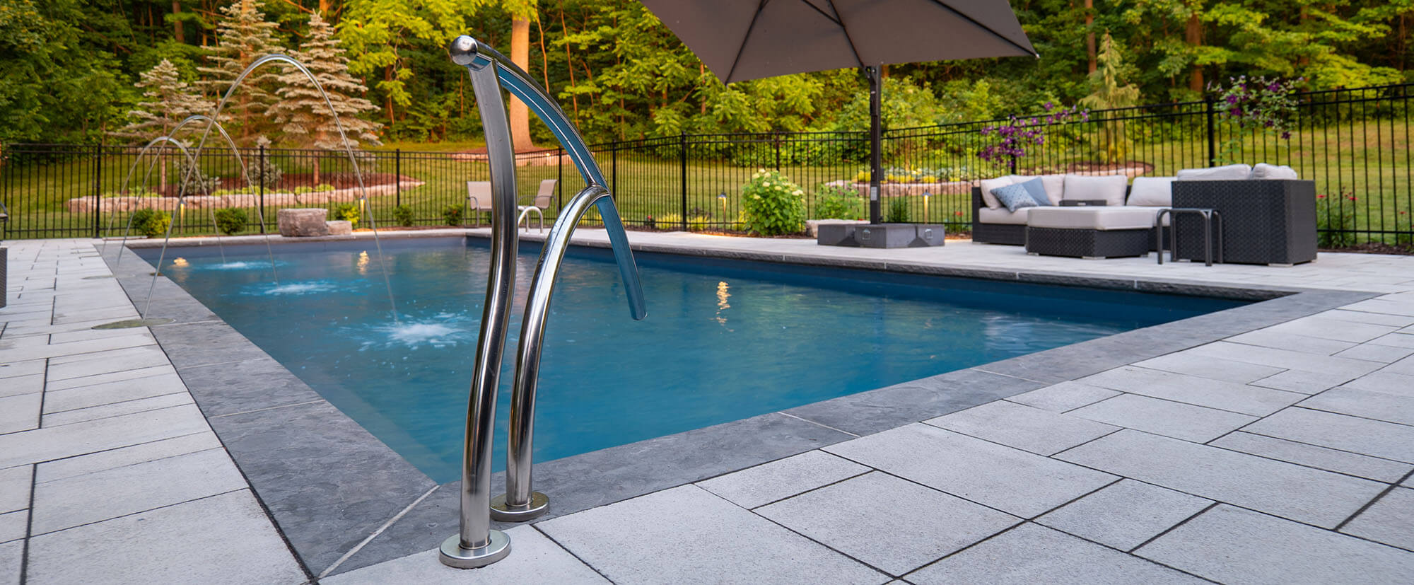 Modern backyard pool with water jets, metal handrail, lounge sofa under umbrella, and green trees beyond fence.