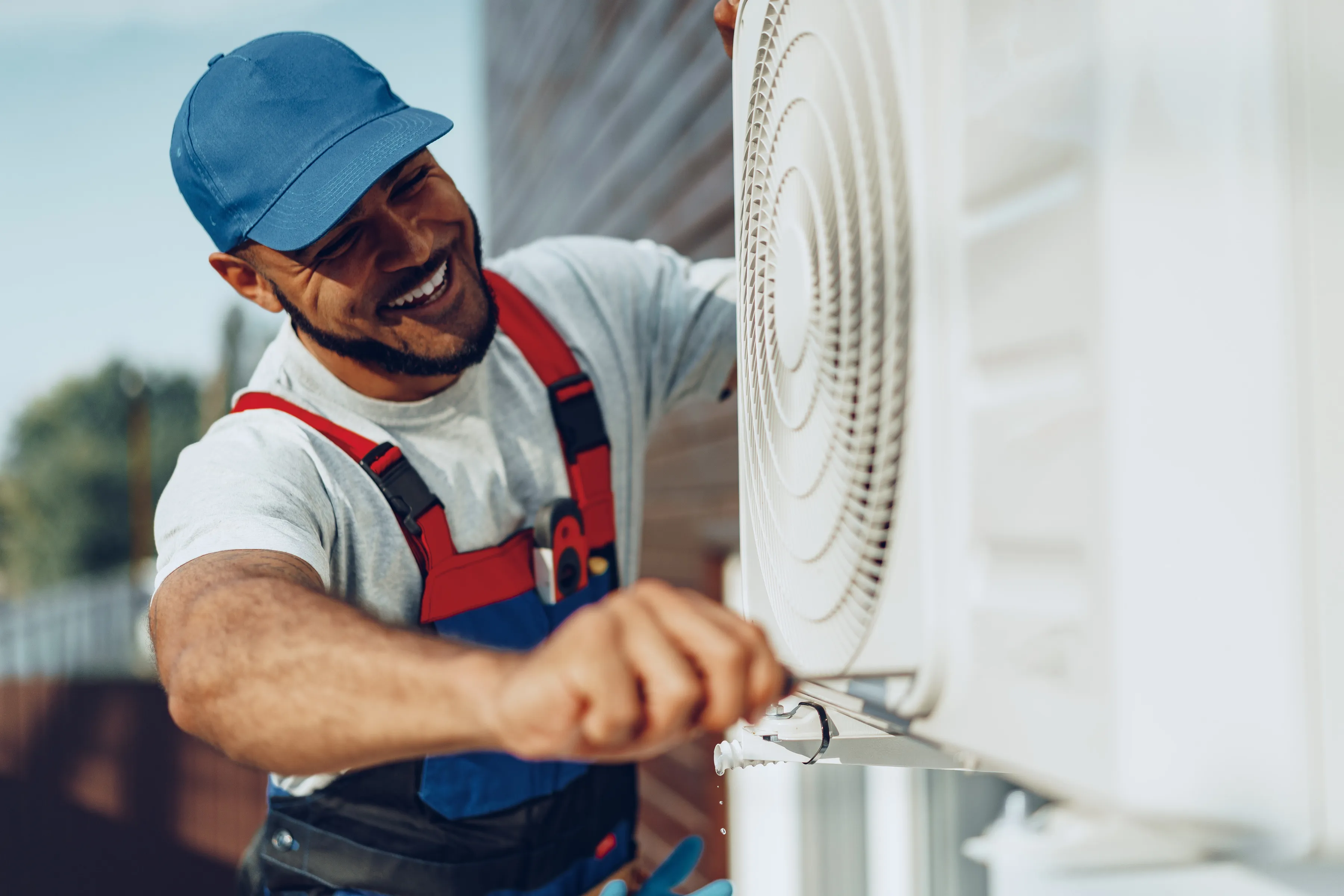 Smiling technician in blue cap and red overalls fixing an outdoor air conditioning unit with a screwdriver.