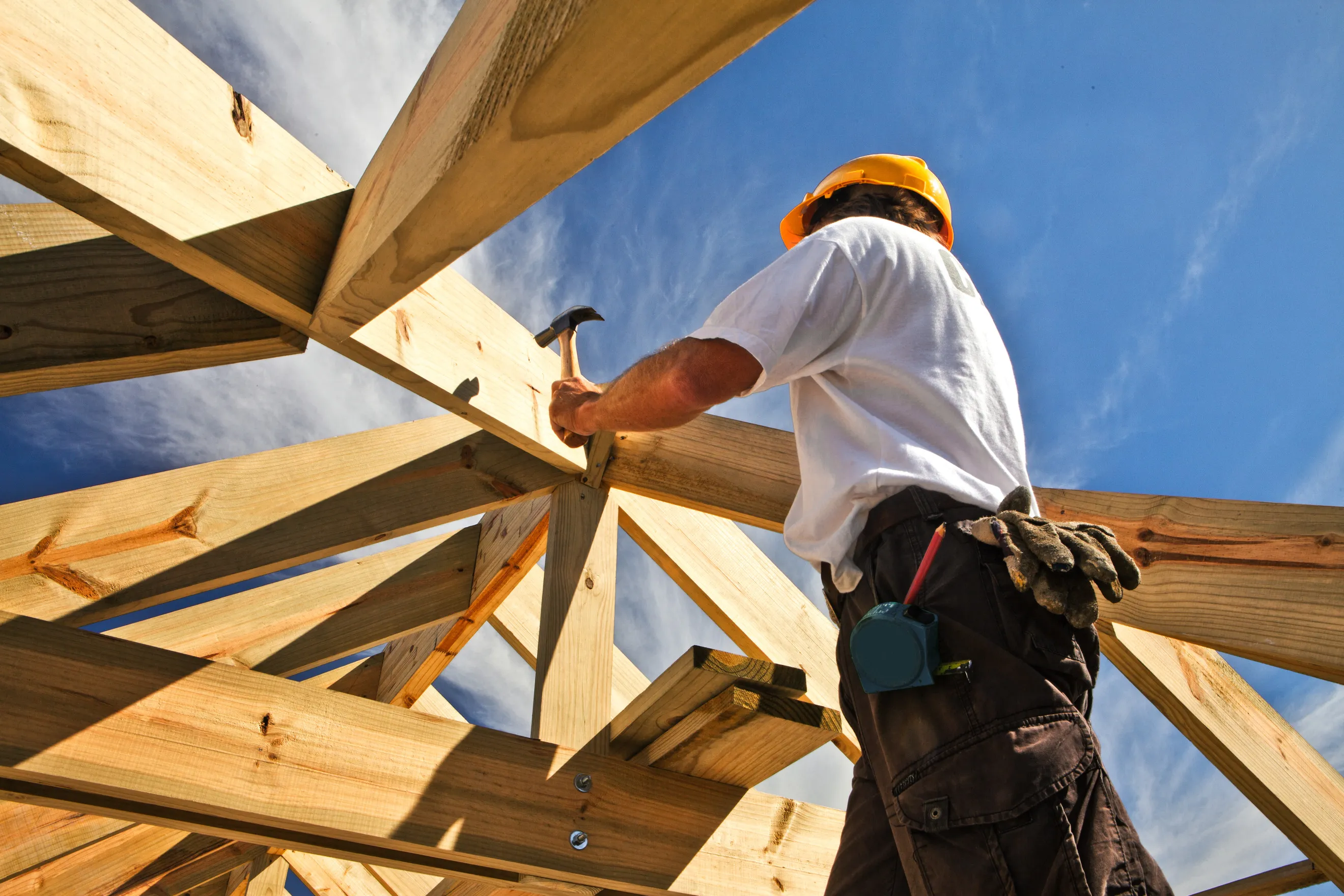 Construction worker in a yellow hard hat using a hammer on wooden roof framing under a blue sky.