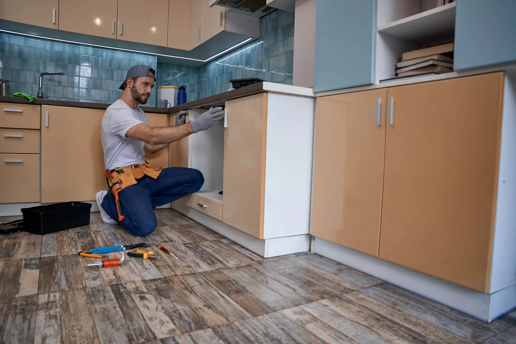 Handyman wearing gloves and a cap kneeling on the floor while repairing a kitchen cabinet door with tools spread on the floor.