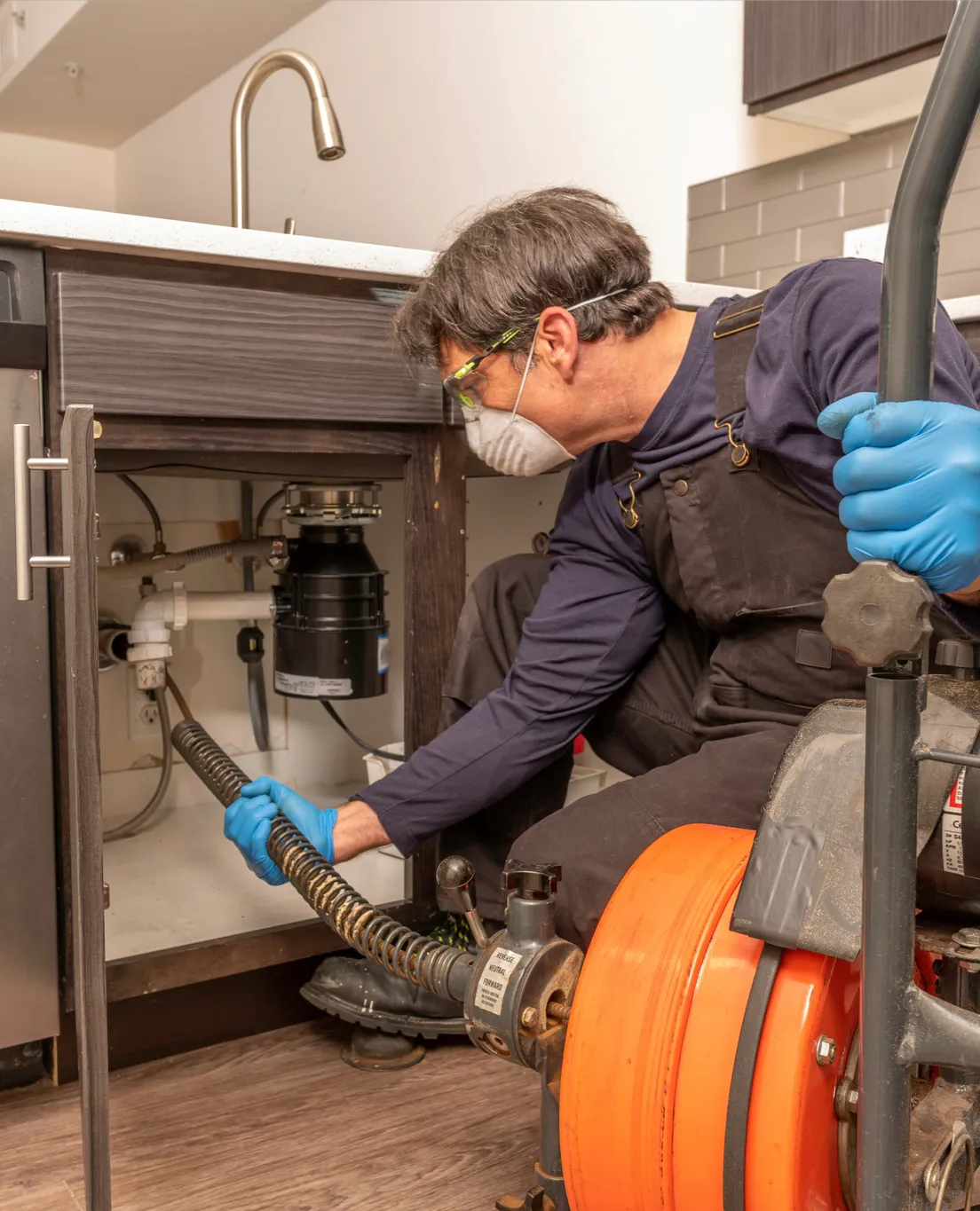 Plumber wearing gloves, goggles, and a mask using a drain cleaning machine under a kitchen sink.
