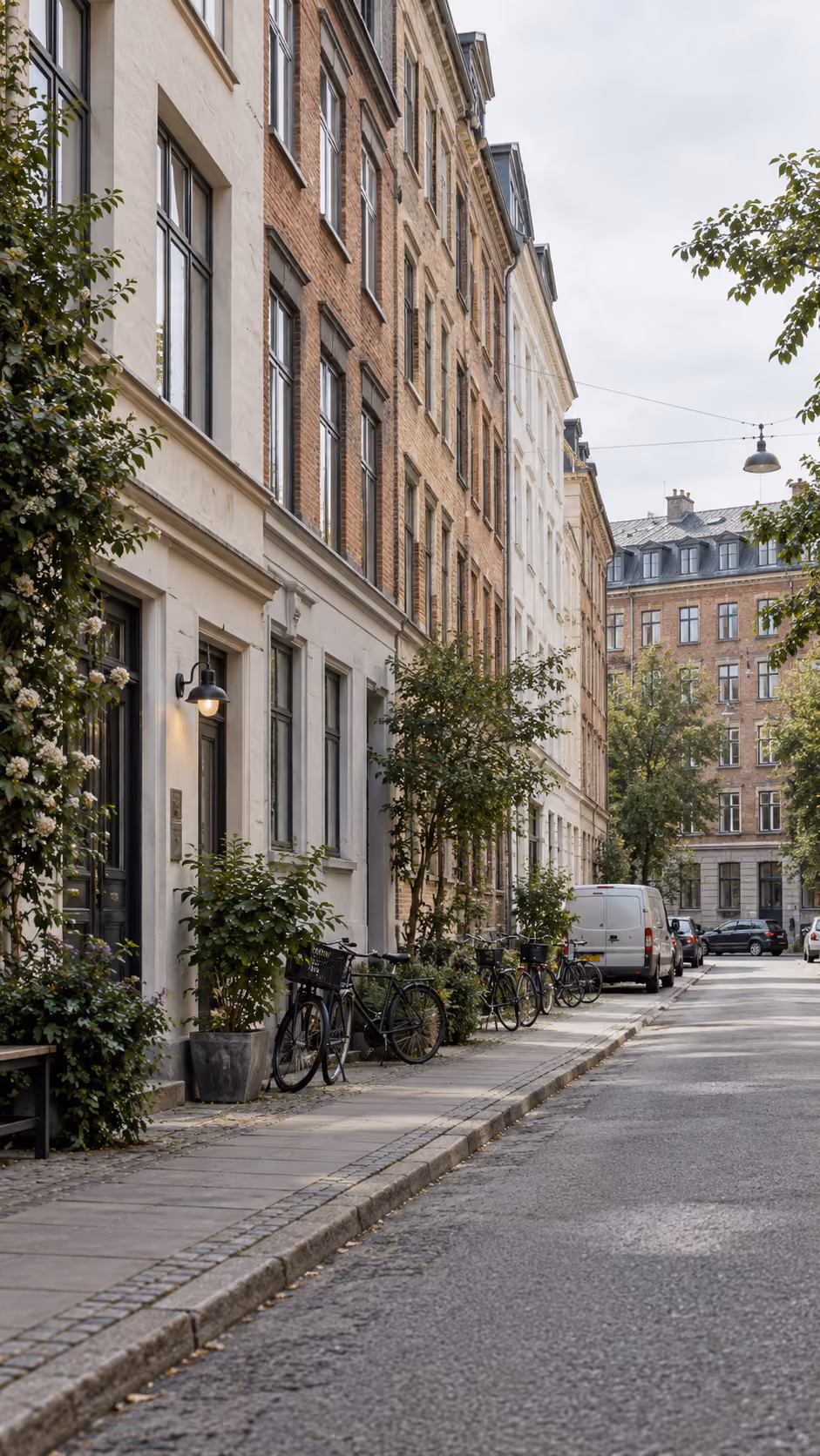 Gade i en by med rækkehuse i mursten og pudsede facader, fortov med cykler parkeret op ad bygningerne og nogle træer langs vejen under en grå himmel.