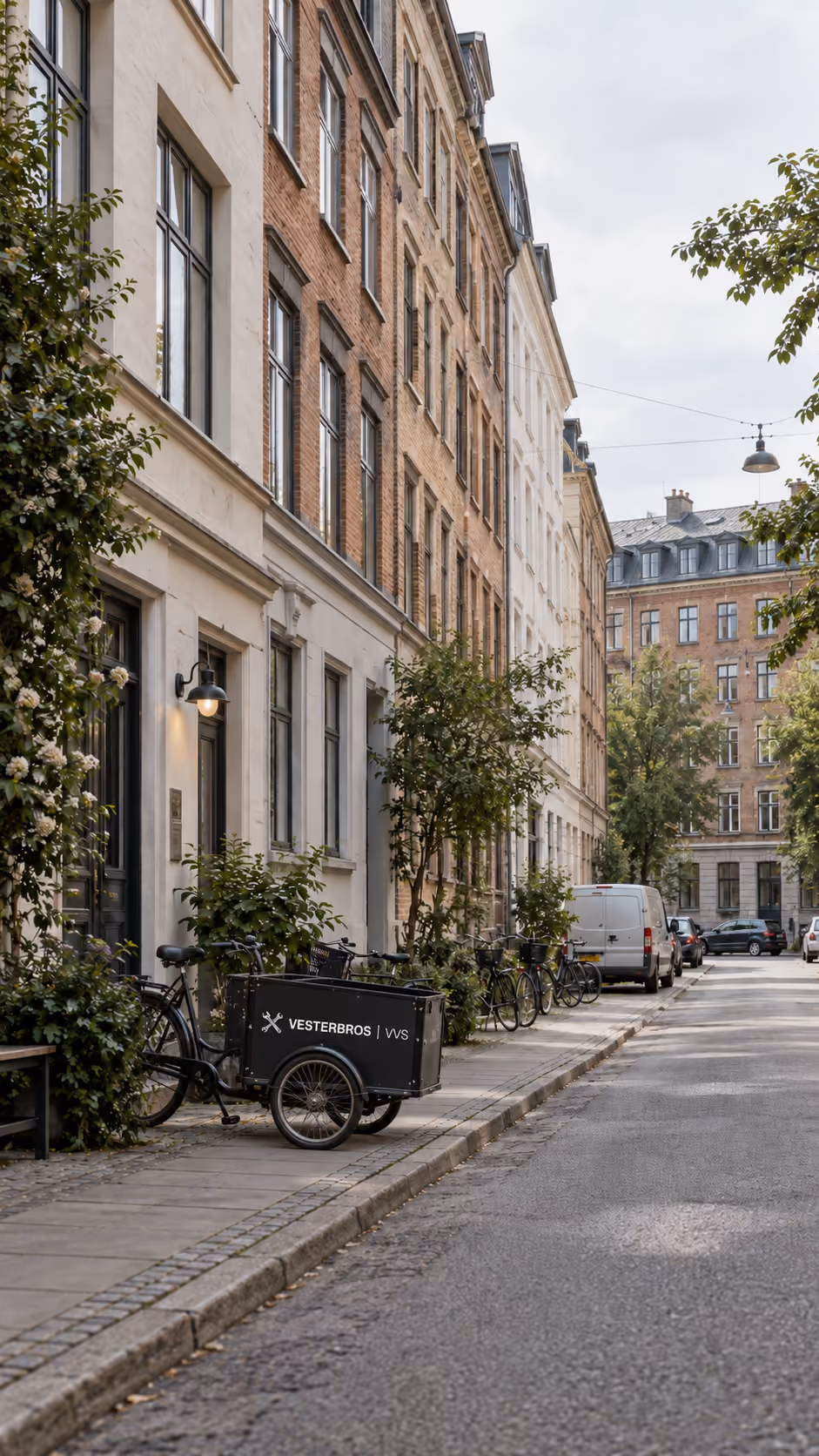 En stille gade med cykler og en varecykel med logoet 'Vesterbros VVS' parkeret ved fortovet foran rækkehuse i en by.