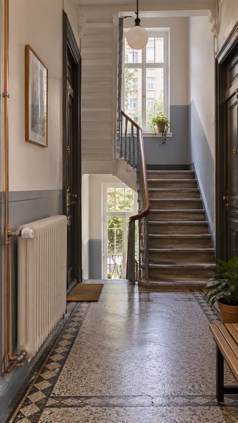 Indendørs trappeopgang med marmortrappe, store vinduer med udsigt til træer, grå og hvide vægge, radiator, planter på gulvet og ved vinduet, og to sorte døre med messinghåndtag.