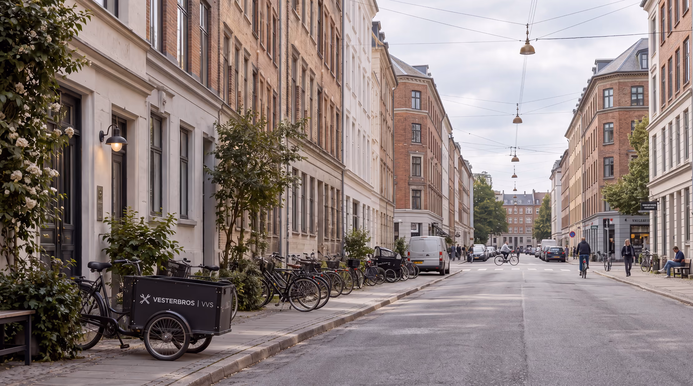 Gadekig i by med cykler parkeret langs fortove på begge sider og personer, der cykler og går under hængende gadelamper mellem fleretagers bygninger.