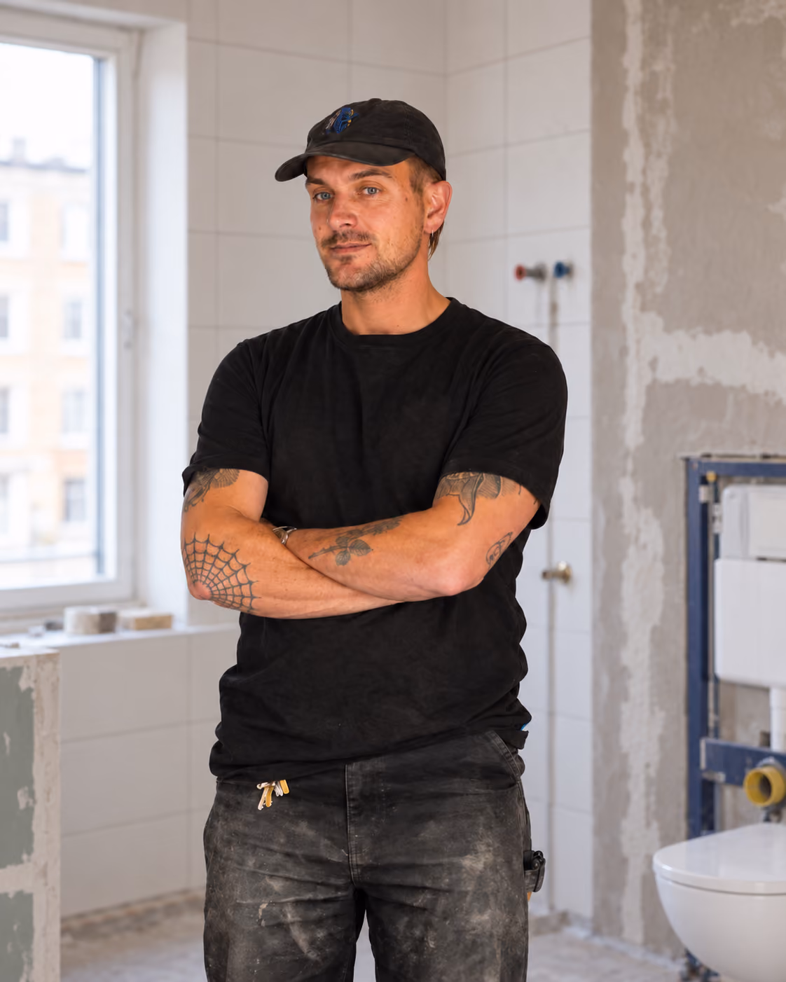 Mand med tatoveringer iført sort kasket, sort t-shirt og støvede bukser står med arme krydsede i et delvist renoveret badeværelse med fliser og toilet.