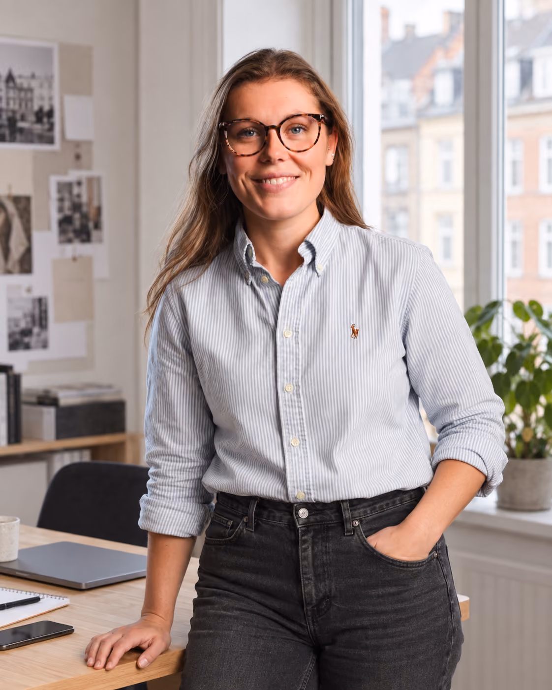 Smilende kvinde med brunt hår, briller og stribet skjorte med ærmerne rullet op, hviler den ene hånd på et skrivebord i et lyst kontor med vinduer og planter bagved.