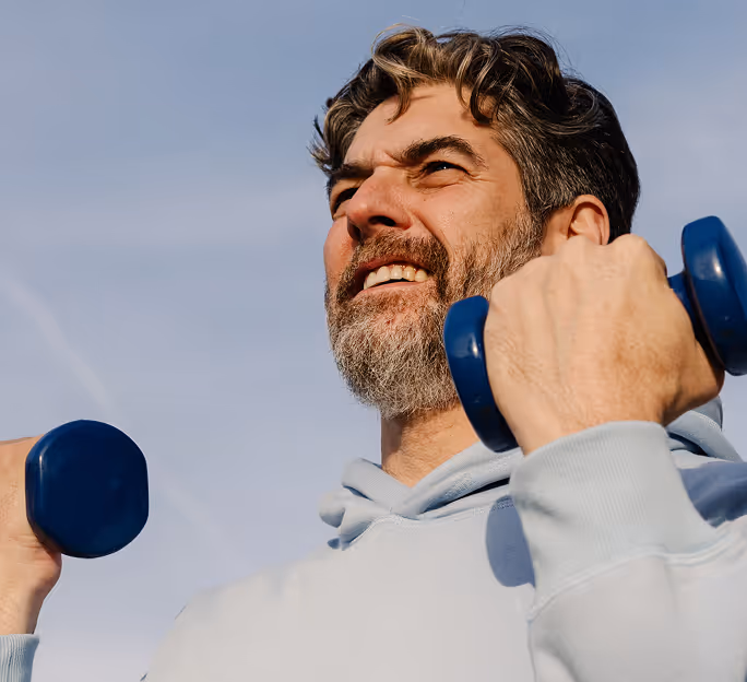 Middle-aged man in a light blue hoodie lifting blue dumbbells outdoors against a clear sky.