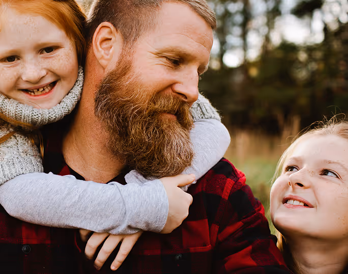 Bearded man in red plaid shirt smiling with two young girls outdoors, one hugging him from behind and the other looking up at him.