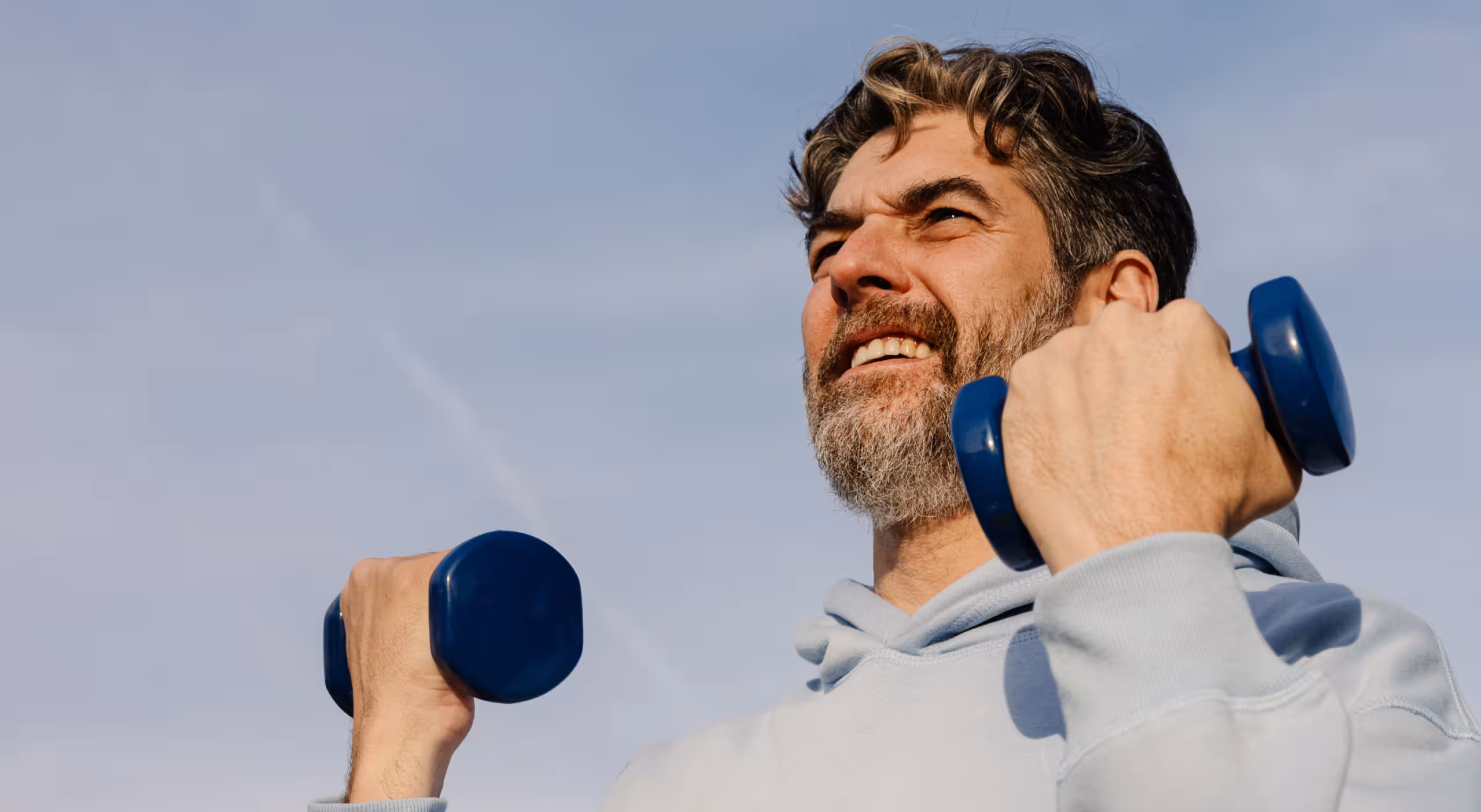 Bearded man in a light hoodie lifting blue dumbbells against a clear sky.