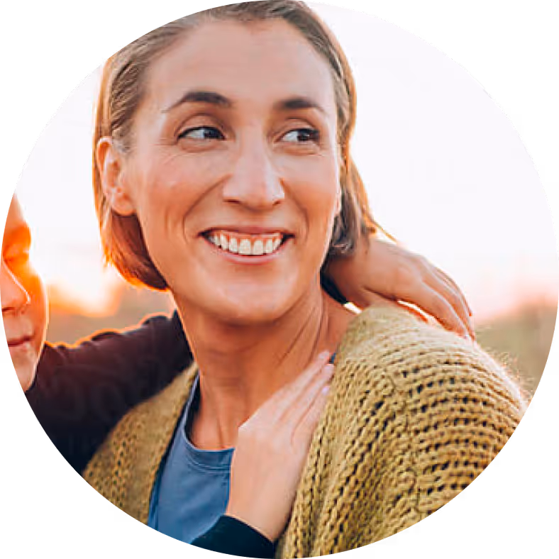 Smiling woman with short hair wearing a knitted sweater, with a child's hand resting on her shoulder.