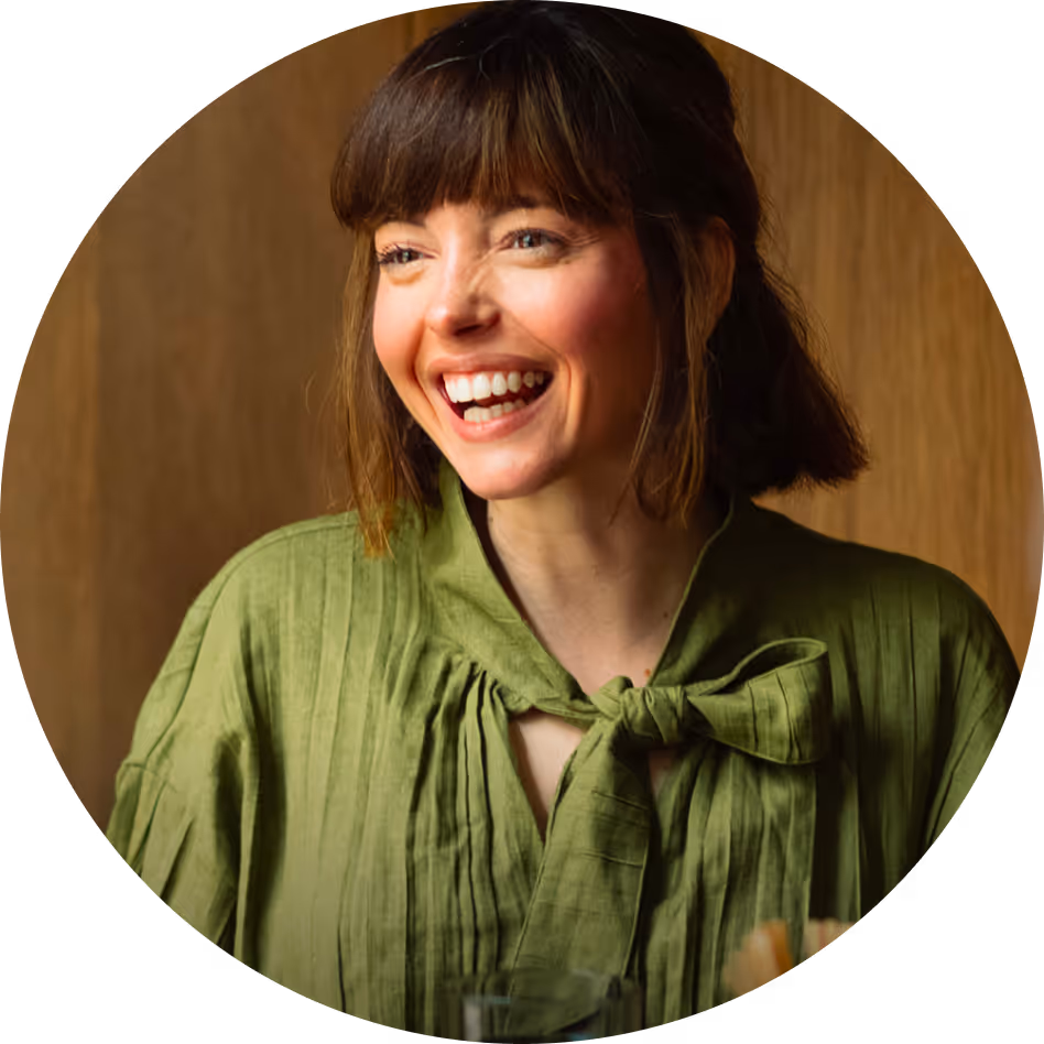 Smiling woman with short brown hair wearing a green blouse with a bow tie, against a wooden background.