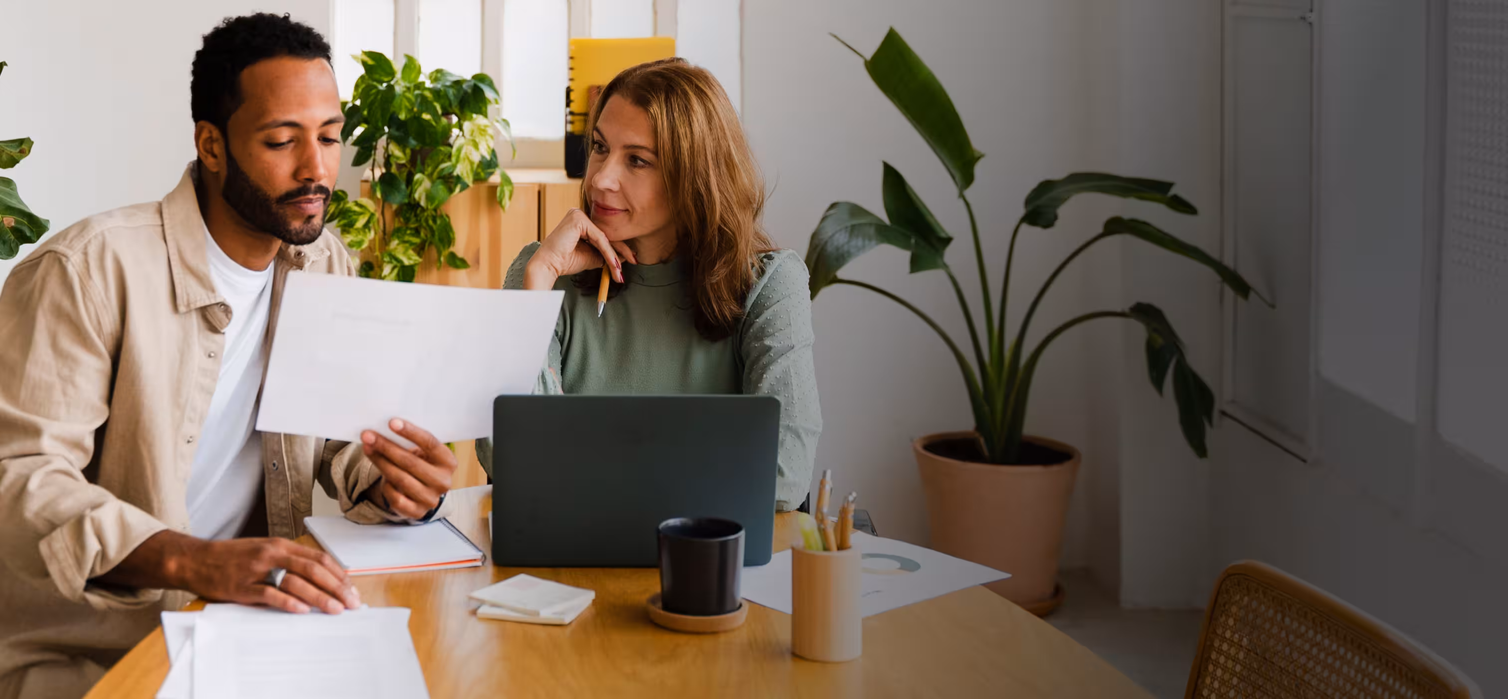 A man and woman sitting at a table reviewing documents with a laptop and plants in the background.