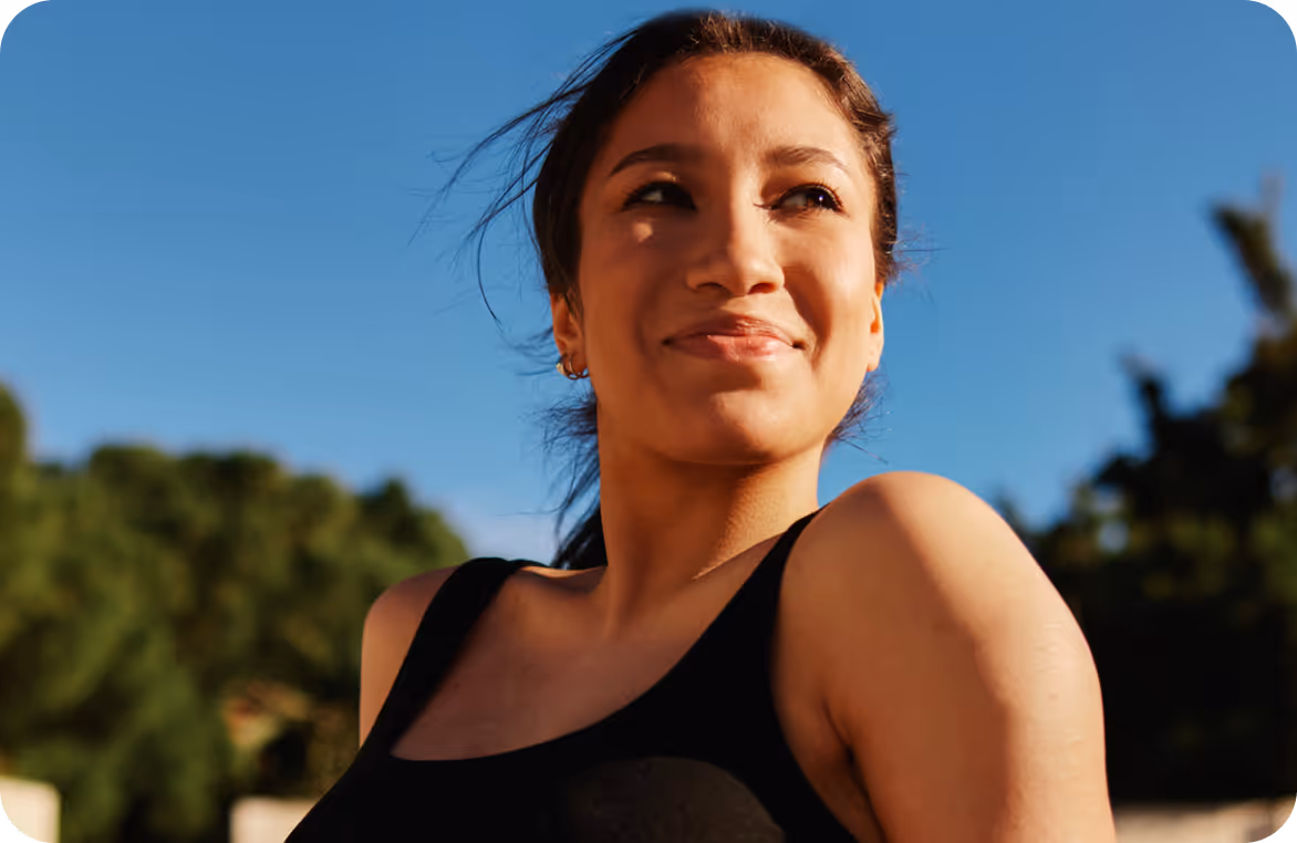 Young woman in a black tank top smiling outdoors with trees and blue sky in the background.