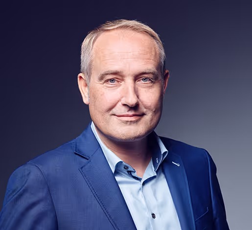 Porträt eines lächelnden Mannes mittleren Alters mit kurzen blonden Haaren in einem blauen Anzug und hellblauem Hemd vor grauem Hintergrund.