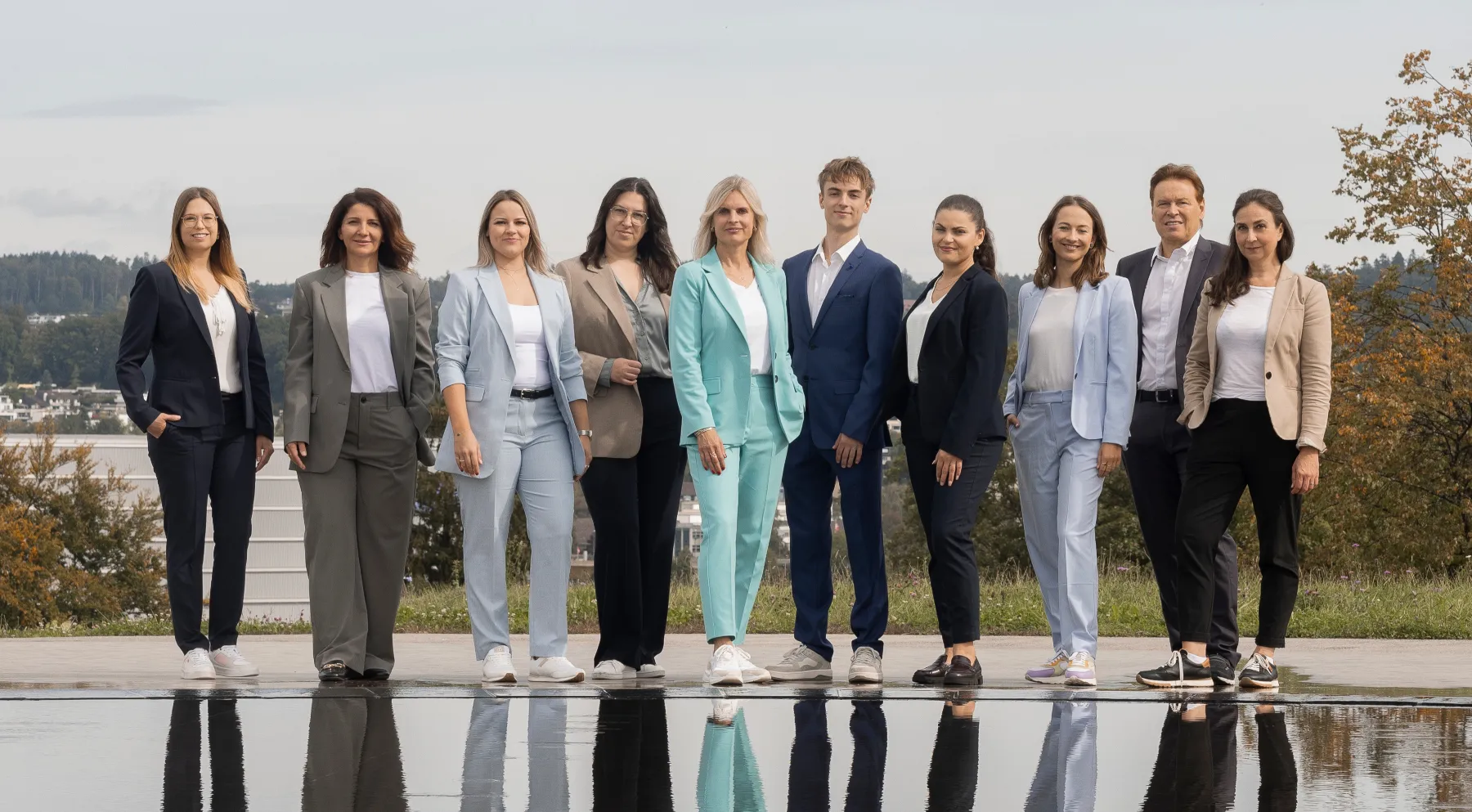 Elf Personen in Businesskleidung stehen nebeneinander draußen vor einem Pool mit Reflexionen und grüner Landschaft im Hintergrund.
