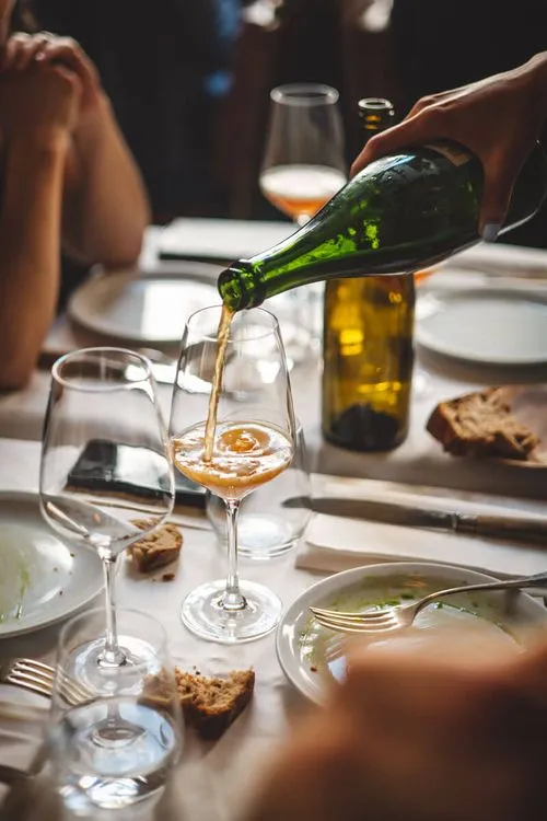 A close-up of a hand pouring an amber-colored natural wine from a green bottle into a glass at a dining table.