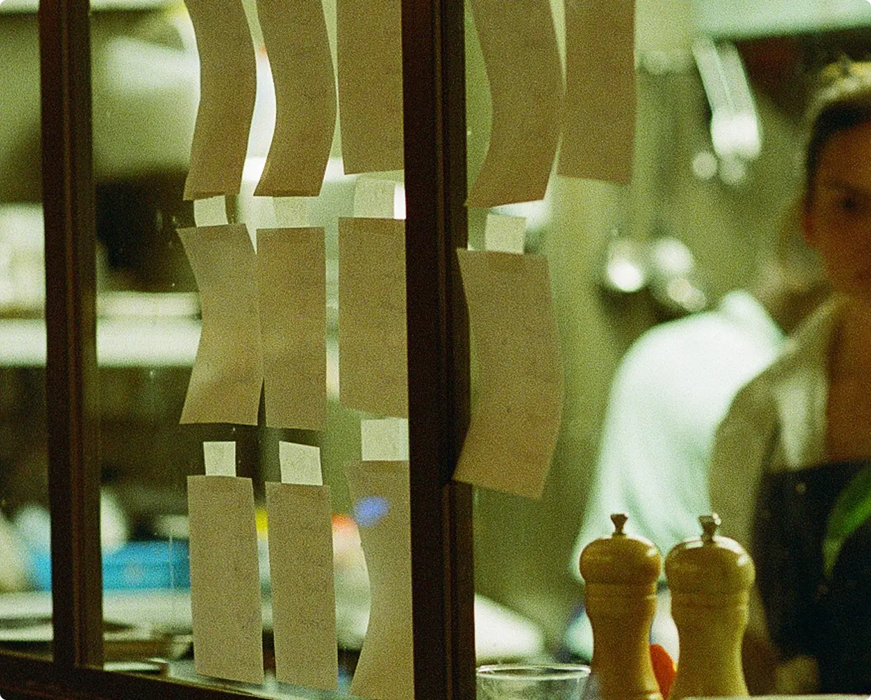 Order tickets hanging in a restaurant kitchen window with a blurred chef in the background.