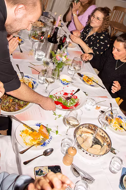 People seated at a table enjoying a meal with wine glasses, plates of food, and a flower centerpiece.