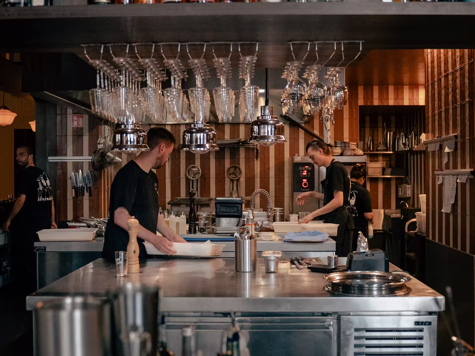 Chefs working in a restaurant kitchen with stainless steel counters and hanging glassware.