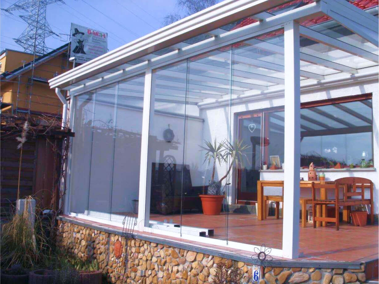 Un adapost pentru masini cu un cadru si acoperis metalic, adiacent unei case cu usi glisante din sticla, inchis de un gard din grilaj metalic. Imaginea prezinta o scena in aer liber cu o structura de adapost pentru masini. Adapostul are un cadru robust de metal gri si este acoperit cu un acoperis alb. O parte din spatele unui vehicul este vizibila in partea stanga sub adapostul pentru masini. Exista un gard din grilaj metalic care inchide zona din jurul adapostului pentru masini, vopsit intr-o culoare inchisa. In spatele gardului, se vede o intrare intr-o casa, caracterizata prin usi glisante din sticla si perdele in interior. Pamantul este pavat cu dale de piatra patrate. Exista cateva plante vizibile in ghivece in spatele gardului.