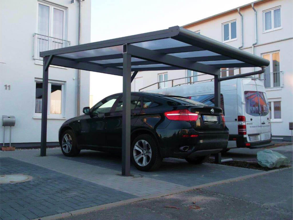 O pergola acoperita atasata unei case galbene cu o banca de lemn, o planta in ghiveci si o alta banca sub adapost. Imaginea prezinta o curte bine intretinuta cu o pergola acoperita atasata unei case. Casa este vopsita in galben deschis si are mai multe usi de sticla care duc spre pergola intrare. O structura metalica albastru inchis sustine acoperisul gri al pergolei intrare. Sub zona acoperita, exista o banca de lemn cu picioare metalice si o alta banca de culoare mai deschisa fara spatar. O planta mare in ghiveci este plasata langa una dintre usile de sticla pe podeaua pergolei intrare placata cu gresie. Curtea are iarba verde tunsa frumos, iar in fundal se poate vedea o parte dintr-o alta cladire.