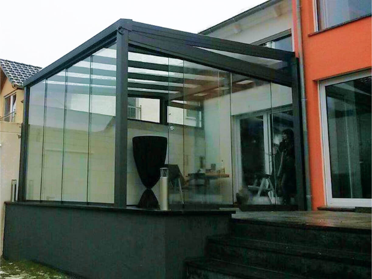 Un carport modern cu un acoperis gri este atasat la o cladire de culoare piersica, cu o poarta de lemn si iarba verde in prim-plan.