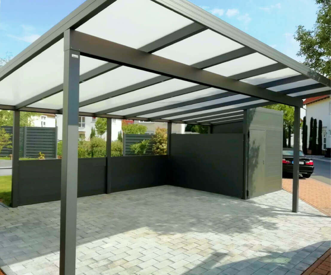 O structura de baldachin metalic atasata lateral unei cladiri albe, oferind adapost peste o zona exterioara pavata. Imaginea prezinta o scena in aer liber cu o structura mare de baldachin metalic atasata lateral unei cladiri. Baldachinul este realizat din grinzi metalice gri si are un acoperis inclinat acoperit cu panouri translucide. Acesta ofera adapost peste o zona pavata cu dale de piatra. In stanga, exista o parte a peretelui exterior alb al unei cladiri, iar pe acesta este montat un felinar de exterior. In fundal, exista tufe verzi care formeaza un gard natural si se poate vedea o alta cladire din caramida rosie in spatele lor.