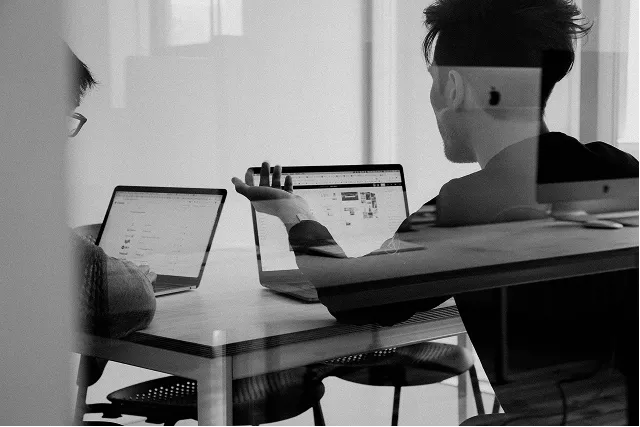 Two people working together at a table with laptops in an office setting.